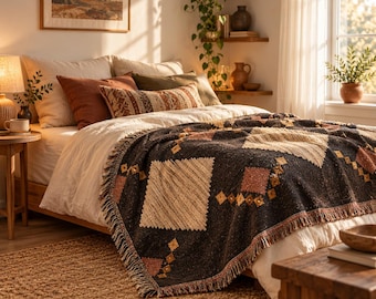 Op de afbeelding: Een geweven plaid met een donkergrijze basis, crème en terracotta ruitpatronen en franjes, gedrapeerd over een bed met gelaagde kussens in tinten crème, roest en olijfgroen in een zonnige slaapkamer.