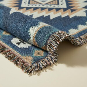 Op de afbeelding: Een geweven plaid met een geometrisch patroon in marineblauw, cr&egrave;me en beige tinten. De stof heeft een getextureerd, gebreid uiterlijk met een omgevouwen hoek die de onderkant laat zien en decoratieve franjes langs de randen.