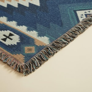 Op de afbeelding: Een geweven plaid met een diepblauwe basis en geometrische patronen in wit, beige en groenblauw. De deken heeft een getextureerd oppervlak en is afgewerkt met decoratieve franjes langs de randen, met een herhalend ruitpatroon.