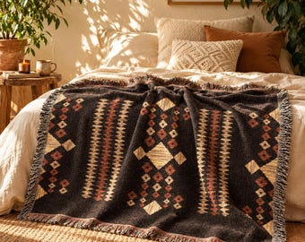 Op de afbeelding: Een donker antracietgrijze geweven plaid met een herhalend geometrisch patroon in tinten crème, roestoranje en bruin. De deken heeft een textuurrijk oppervlak met een franjerand en ligt over een bed met kussens in neutrale kleuren.