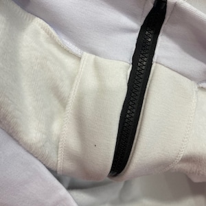 May include: Close-up of a white zipper on a white garment. The zipper is black and has a metal slider.