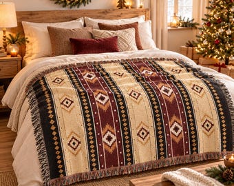 Op de afbeelding: Een geweven plaid met een herhalend geometrisch patroon in de kleuren crème, donkerrood, zwart en beige. De deken heeft een rand met franjes en ligt over een bed met wit beddengoed, aangevuld met diverse decoratieve kussens.