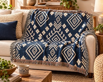 Op de afbeelding: Een geweven plaid in marineblauw en crème met een herhalend geometrisch ruitpatroon en franjes aan de randen, gedrapeerd over een lichtgekleurde bank. Het textiel vertoont een contrastrijk ontwerp met scherpe lijnen en kleine vierkantjes.