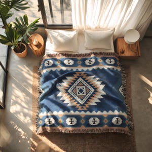 Op de afbeelding: Een geweven plaid met een diepblauwe achtergrond, een groot centraal ruitpatroon in wit en beige, geaccentueerd door kleinere geometrische vormen en een franjerand, gedrapeerd over een bed met twee witte kussens.