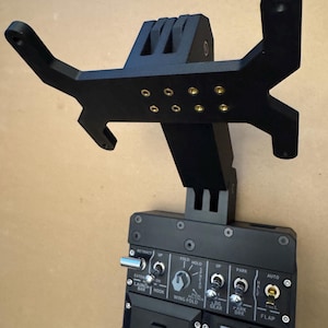 Könnte beinhalten: Eine schwarze Steuertafel mit verschiedenen Schaltern, Drehreglern und Beschriftungen, darunter "RETRACT", "FOLD HOLD" und "FLAP". Eine schwarze Halterung ist oberhalb der Tafel angebracht und weist mehrere Befestigungslöcher und eine markante Form auf. Die Tafel hat einen roten Knopf und einen Schieberegler.