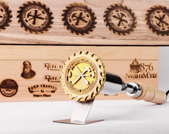 May include: A wood-burning stamp with a circular design featuring a hammer and cross symbol. The stamp is mounted on a metal stand with a wooden handle. The background includes wooden signs with similar designs and text.