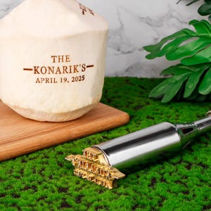 Könnte beinhalten: Eine weiße Kokosnuss mit dem Text "THE KONARIK'S - APRIL 19, 2025" eingraviert. Ein Werkzeug aus Metall mit einem Stempel aus Messing, auf dem "THE KONARIK'S" steht, liegt auf einer grünen Oberfläche.