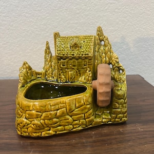 May include: A green ceramic planter shaped like a cottage with a water wheel. The planter has a small opening for planting and is decorated with a stone-like texture.