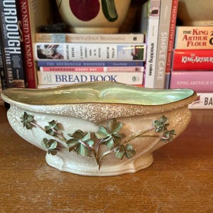 May include: Cream-colored ceramic bowl with a light green interior and gold trim. The bowl is decorated with raised green shamrock designs. The bowl is sitting on a wooden surface, with books and other ceramic bowls in the background.