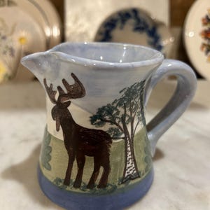 May include: A small, hand-painted ceramic creamer with a blue rim and handle. The creamer features a brown moose silhouette with a tree and landscape design. The background is light blue and green.
