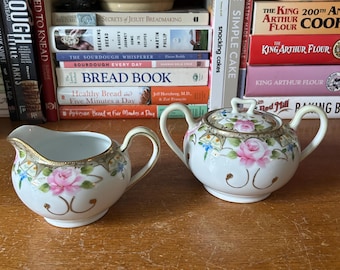 May include: A white porcelain creamer and sugar bowl set. The set features pink roses, gold trim, and delicate blue accents. The creamer has a handle and spout, while the sugar bowl has a lid and handles. The set is placed on a wooden surface.