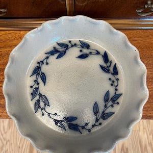 May include: A round, speckled light gray ceramic pie dish with a scalloped edge. The interior features a hand-painted dark blue vine pattern arranged in a circular wreath design, consisting of small leaves and berries painted on the glazed surface.