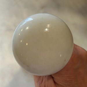 May include: A round, smooth, white ceramic knob with a glossy finish. The surface of the knob is opaque and reflects light, showing a uniform, spherical shape. The material appears to be solid, polished porcelain or ceramic, featuring a clean, neutral aesthetic.