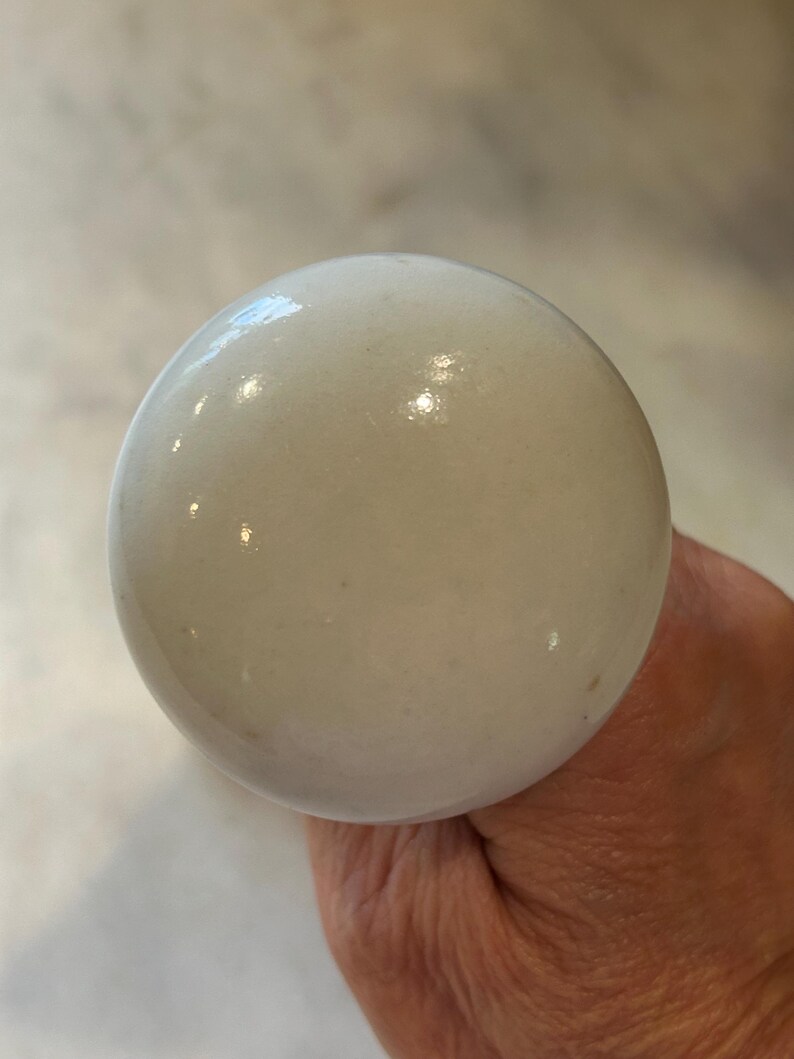 May include: A round, smooth, white ceramic knob with a glossy finish. The surface of the knob is opaque and reflects light, showing a uniform, spherical shape. The material appears to be solid, polished porcelain or ceramic, featuring a clean, neutral aesthetic.