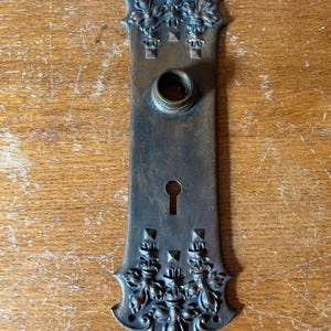 May include: An antique bronze door plate with ornate floral and leaf designs. The rectangular plate features a keyhole and a circular opening for the doorknob. The top and bottom edges have decorative flourishes.