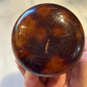 May include: A round, tortoiseshell-patterned object with a glossy finish. The object has a dark brown base with lighter brown and amber marbling. The surface shows minor scratches. The object is likely a decorative item or part of a larger piece.