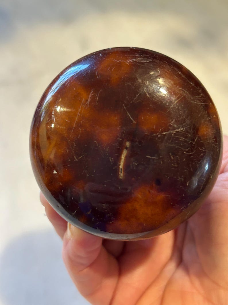 May include: A round, tortoiseshell-patterned object with a glossy finish. The object has a dark brown base with lighter brown and amber marbling. The surface shows minor scratches. The object is likely a decorative item or part of a larger piece.