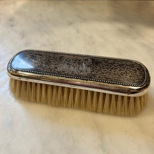 May include: A vintage silver-toned brush with a rectangular shape. The brush features a textured, dark-colored top with an ornate monogram. The bristles are light brown. The brush has a silver-colored trim.