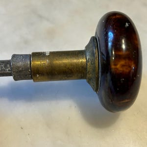 May include: Close-up of a vintage door knob. The knob has a dark brown, oval-shaped handle with a mottled pattern. The handle is attached to a brass and metal mechanism.