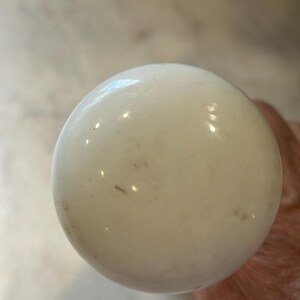 May include: A spherical white ceramic knob with a smooth, glossy finish. The surface of the knob displays subtle, light brown speckles and minor imperfections, reflecting light across its rounded, polished exterior structure.