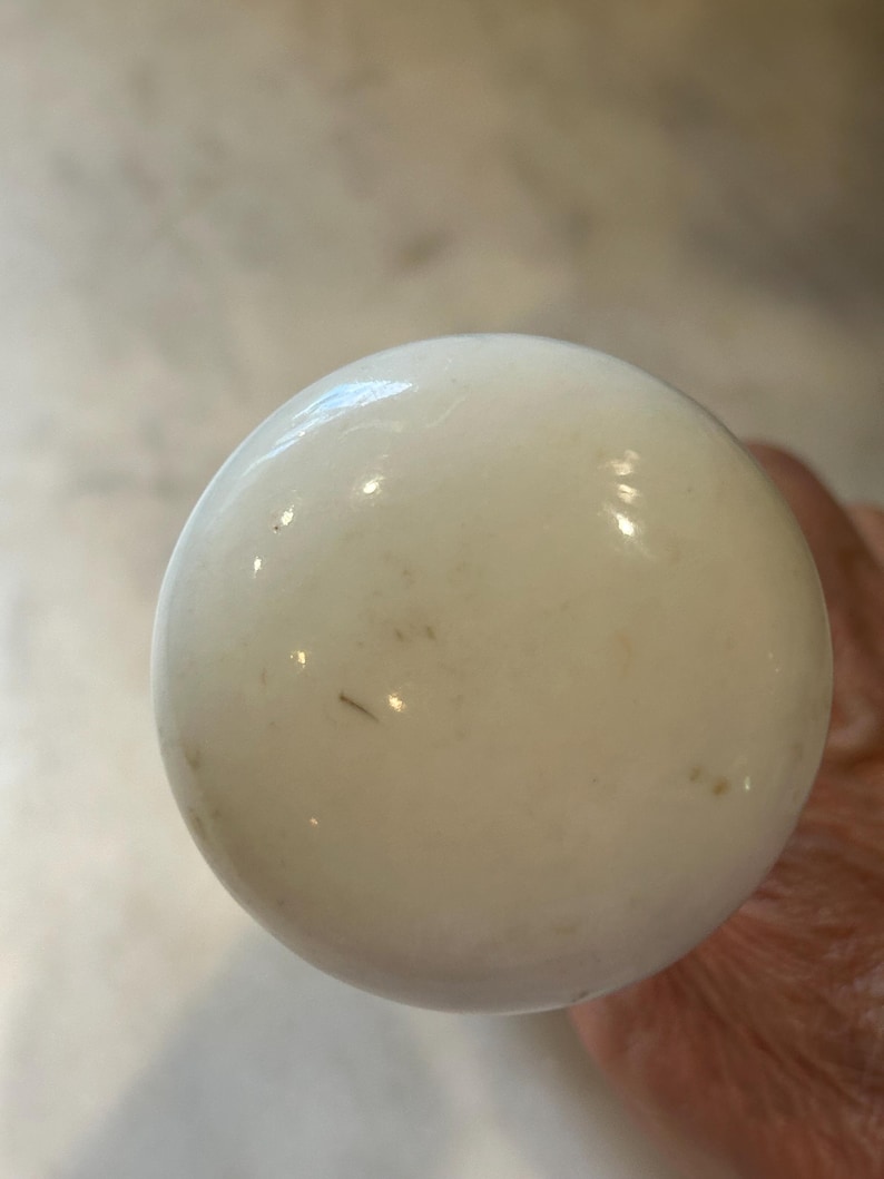 May include: A spherical white ceramic knob with a smooth, glossy finish. The surface of the knob displays subtle, light brown speckles and minor imperfections, reflecting light across its rounded, polished exterior structure.