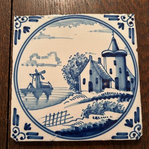 May include: A square ceramic tile featuring a hand-painted blue and white scene of a Dutch landscape. The design includes a windmill, a house, and a tower enclosed within a circular border, with decorative corner motifs on a white background.