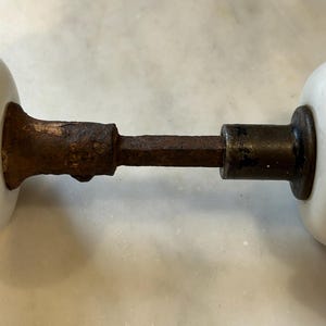 May include: A pair of white porcelain door knobs connected by a central square metal spindle. The metal components exhibit significant surface rust and oxidation, showing a weathered texture across the connecting rod and the base of the knobs.