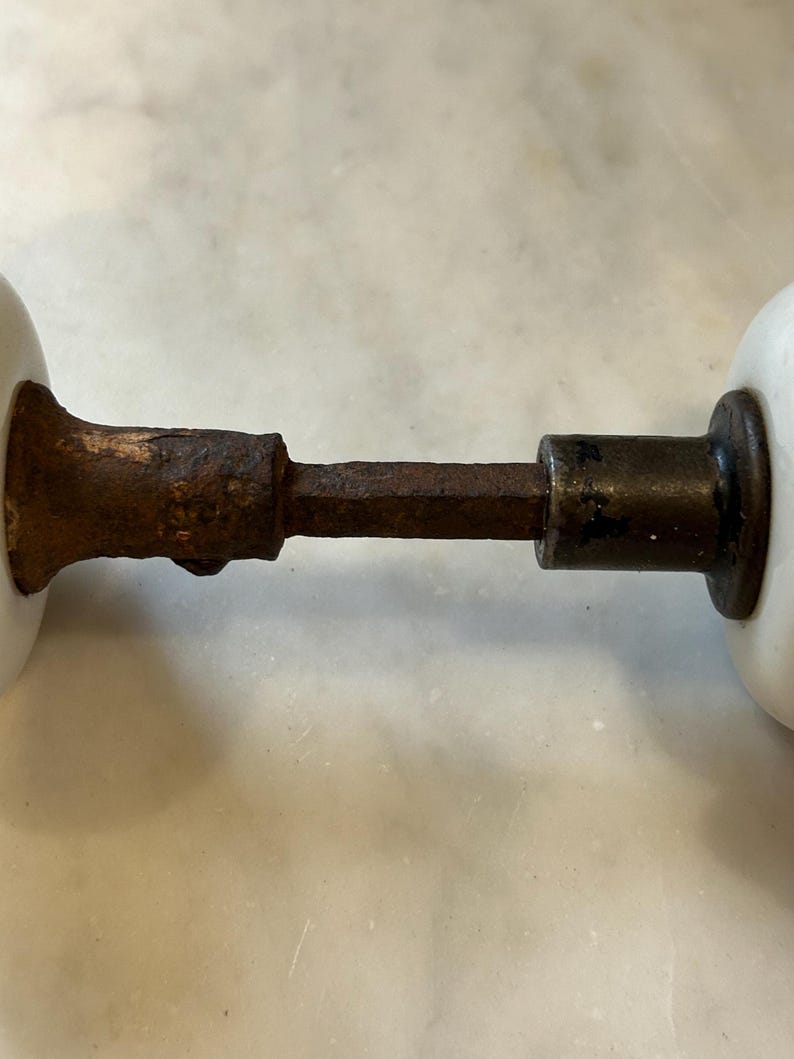 May include: A pair of white porcelain door knobs connected by a central square metal spindle. The metal components exhibit significant surface rust and oxidation, showing a weathered texture across the connecting rod and the base of the knobs.