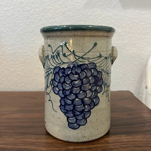 May include: A white ceramic utensil holder with a blue and green grape design. The holder has two handles on the sides.