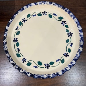May include: A white ceramic pie plate with a blue and green floral design. The plate has a scalloped edge and a speckled blue glaze.