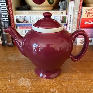 May include: A maroon ceramic teapot with a white lid and a white band around the top. The teapot has a rounded body and a spout on one side and a handle on the other.