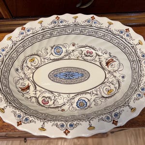 May include: Oval serving platter with a cream-colored base and a decorative floral and scroll design. The rim has a scalloped edge with a repeating pattern of blue, yellow, and red accents. The center features a blue floral design.