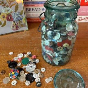 May include: A glass jar filled with a variety of colorful buttons. The jar has a green lid and a wire handle. The buttons are scattered on a wooden surface.