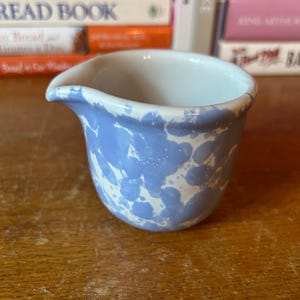 May include: A small, ceramic creamer with a light blue and white splatter pattern. The creamer has a spout and a rounded body. The interior is white. The creamer sits on a wooden surface.