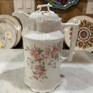 May include: White porcelain pitcher with a lid, featuring a floral design in pink, yellow, and blue. The pitcher has a gold trim and a decorative handle. The design includes embossed details and a scalloped base.