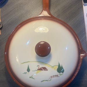 May include: A ceramic skillet with a lid. The skillet has a brown handle and rim, with a white interior. The lid features a brown knob and a painted landscape scene with a house, trees, and a fence.