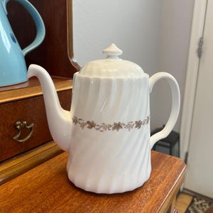May include: White ceramic coffee pot with a brown floral design around the middle. The pot has a swirl design and a lid with a knob.