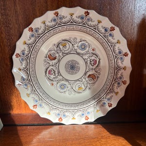 May include: A white ceramic plate with a scalloped edge. The plate features a detailed design with floral and geometric patterns in blue, orange, yellow, and black. The center has a circular design with a star-like pattern.