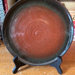 May include: A handmade ceramic plate with a spiral design in the center. The plate features a reddish-brown interior with a dark green rim. It is displayed on a black wooden stand, showcasing its unique craftsmanship.