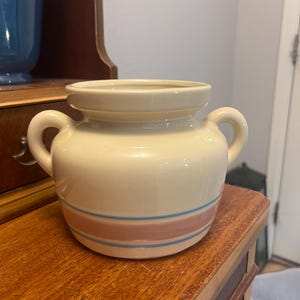May include: Cream-colored ceramic jar with two handles and a wide opening. The jar features two horizontal stripes, one pink and one blue, around the middle. The jar is sitting on a wooden surface.