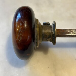 May include: Close-up of a vintage door knob. The knob is oval-shaped and made of a dark brown, glossy material. It has a brass-colored base and a metal rod extending from the back. The knob shows signs of age and wear.