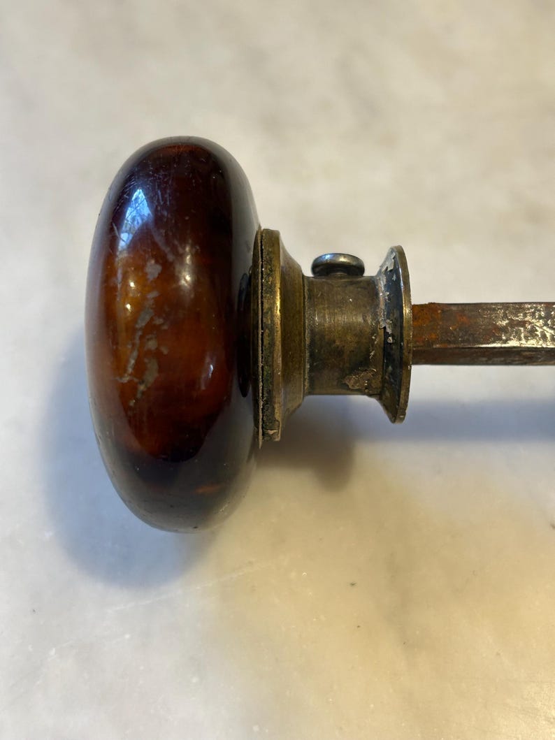 May include: Close-up of a vintage door knob. The knob is oval-shaped and made of a dark brown, glossy material. It has a brass-colored base and a metal rod extending from the back. The knob shows signs of age and wear.