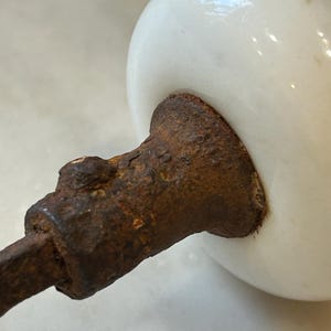 May include: A round, white ceramic knob attached to a heavily rusted, textured metal spindle. The white surface of the knob is smooth and glossy, contrasting with the dark, corroded, and pitted appearance of the metal hardware component.