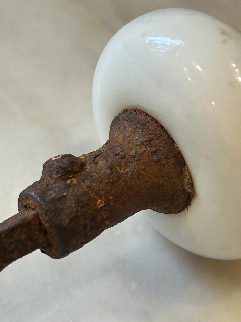 May include: A round, white ceramic knob attached to a heavily rusted, textured metal spindle. The white surface of the knob is smooth and glossy, contrasting with the dark, corroded, and pitted appearance of the metal hardware component.