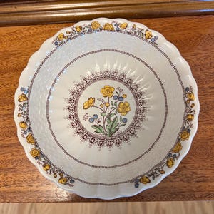 May include: A round ceramic bowl with a scalloped edge, featuring a cream-colored base with a textured basket-weave pattern. The center displays a floral bouquet of yellow and blue flowers, surrounded by a brown decorative border and floral rim.