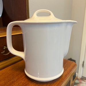 May include: A white ceramic teapot with a lid and a handle. The teapot is sitting on a wooden surface.