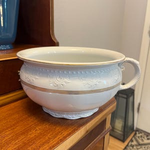 May include: A white ceramic chamber pot with a handle, featuring embossed floral patterns and gold trim. The antique-style pot sits on a wooden surface, showcasing its decorative design and vintage aesthetic.