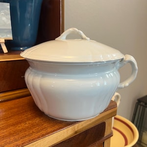 May include: A white ceramic chamber pot with a matching lid, featuring a rounded body with vertical fluted detailing and a single side handle. The lid has a central decorative knob handle, and the entire piece has a smooth, glossy finish.