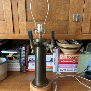 May include: A vintage-style lamp with a brass finish, featuring two tap handles and a wooden base. The lamp has a cylindrical body and a light bulb socket. The lamp is on a wooden surface.