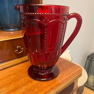 May include: A ruby red glass pitcher with a decorative design. The pitcher has a handle and a spout for pouring. The design includes beaded accents and a textured pattern. It is sitting on a wooden surface.